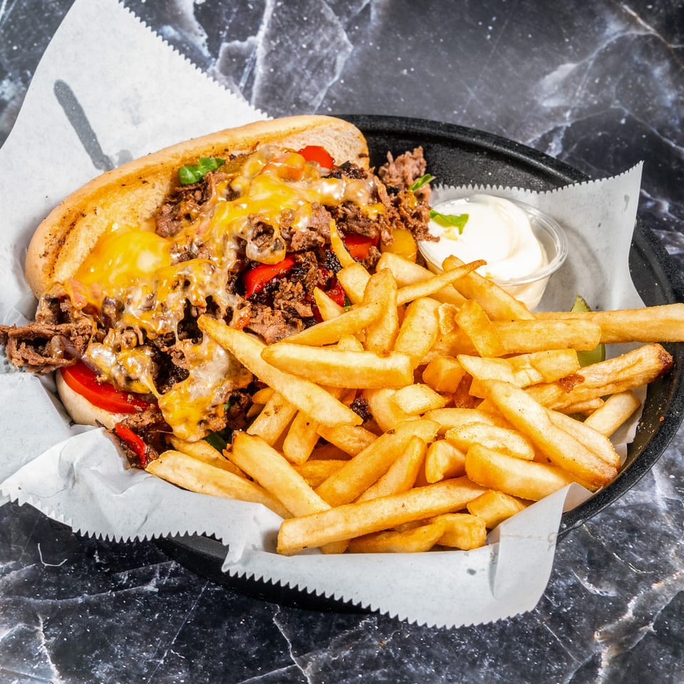 Cheesesteak sandwich with peppers and onions, served alongside golden fries and creamy dipping sauce on a lined tray.
