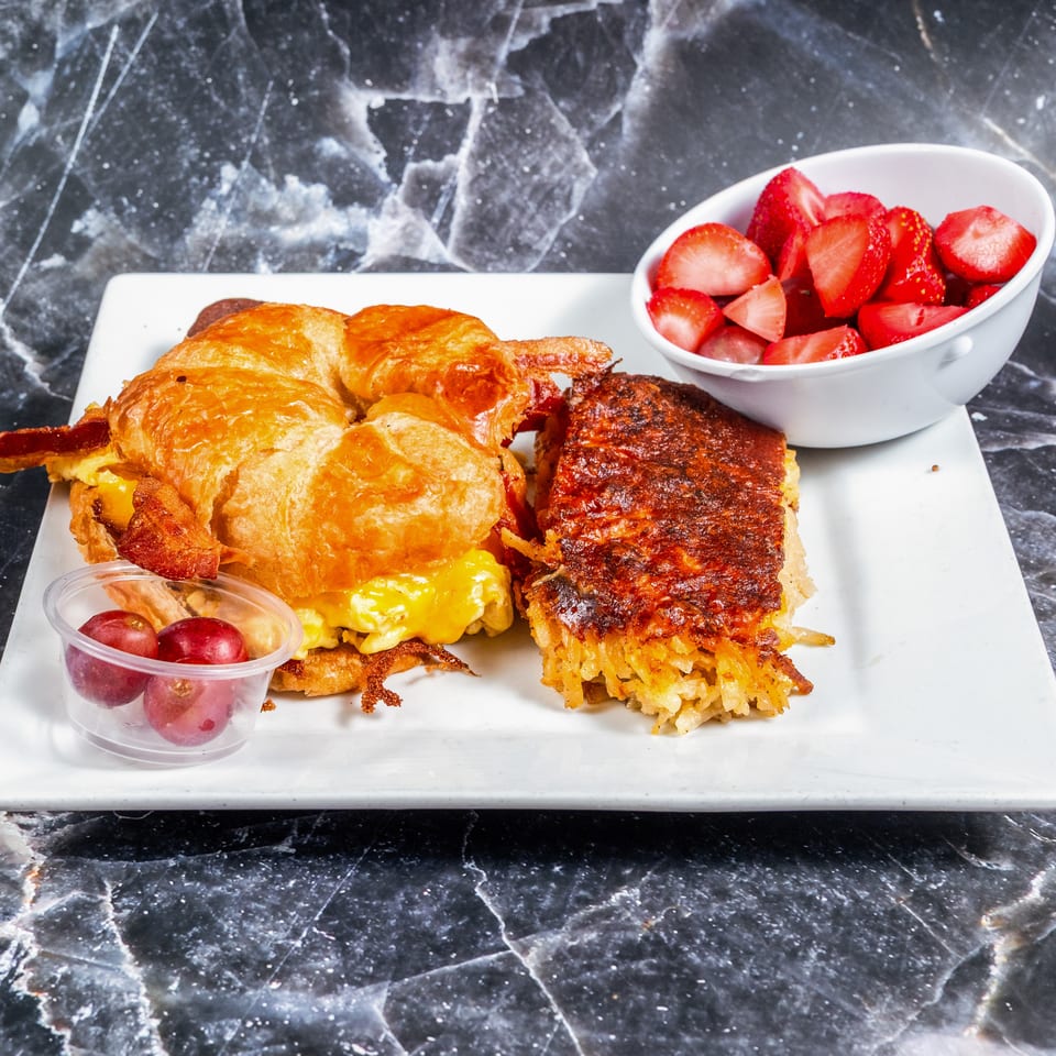 Croissant breakfast sandwiches with bacon, egg, and cheese, served with bowls of strawberries and grapes on a white platter.