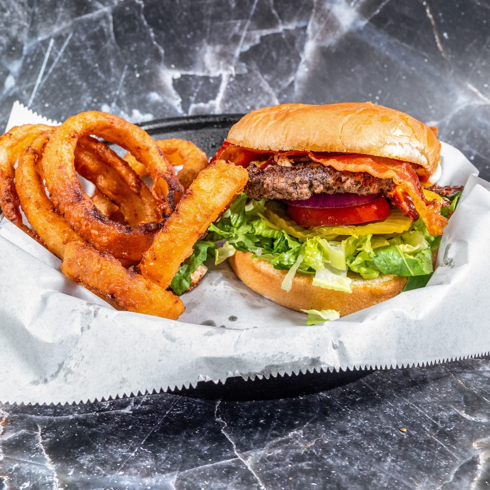 Bacon cheeseburger with lettuce, tomato, pickles, and onions served alongside crispy onion rings on a paper-lined tray.