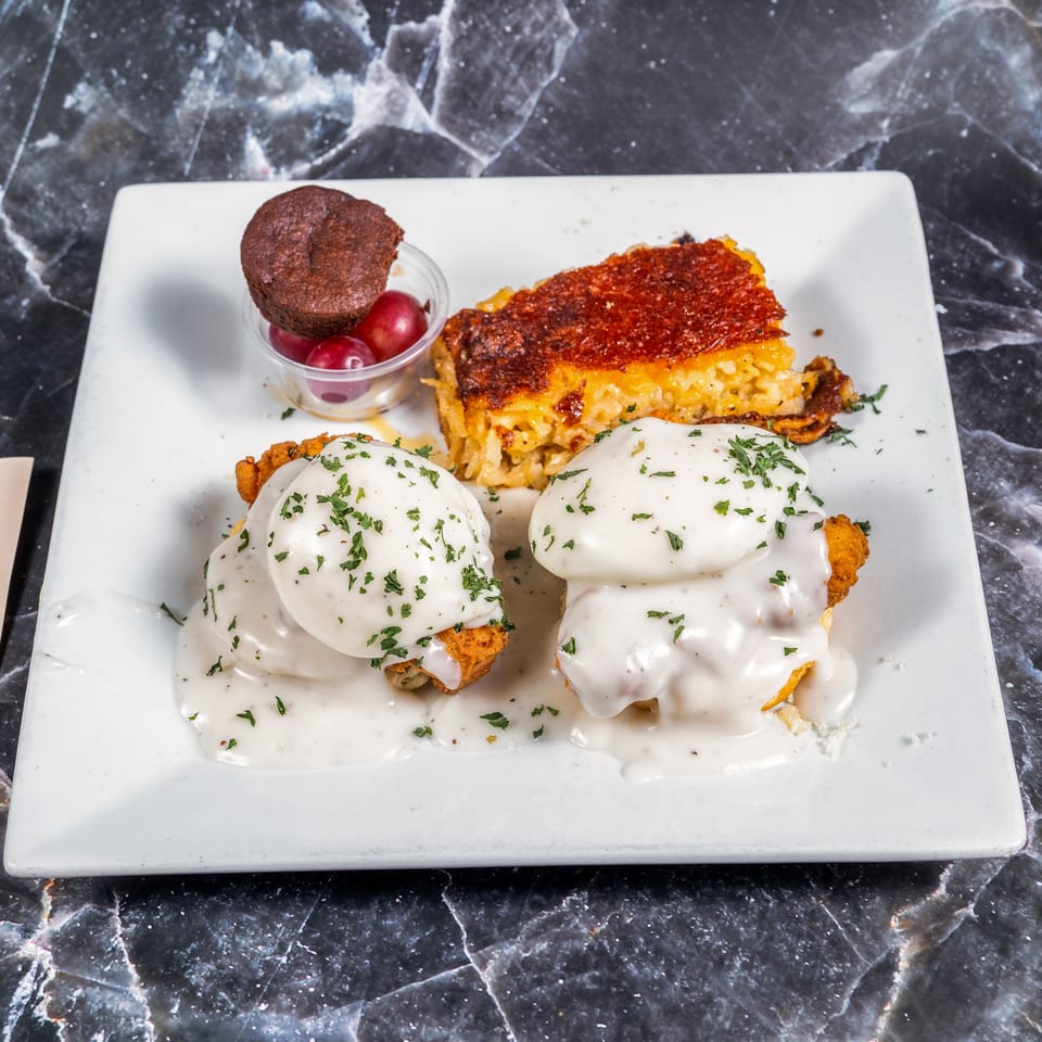 White plate holding two chicken-and-waffle portions with creamy sauce, cheesy baked macaroni, chocolate muffin, and cherries.