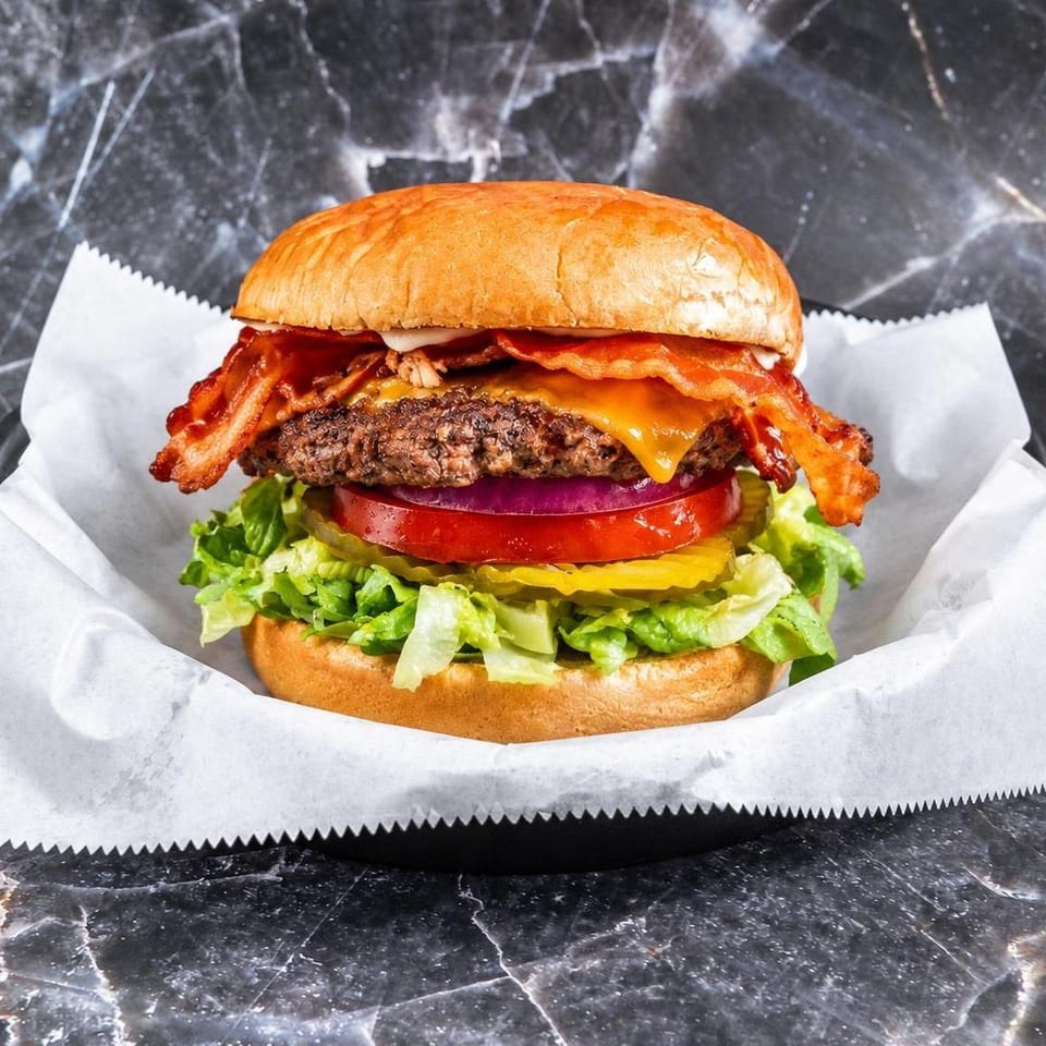 Loaded bacon cheeseburger with lettuce, tomato, pickles, red onion, and sauce, served on white paper over a dark marble table.