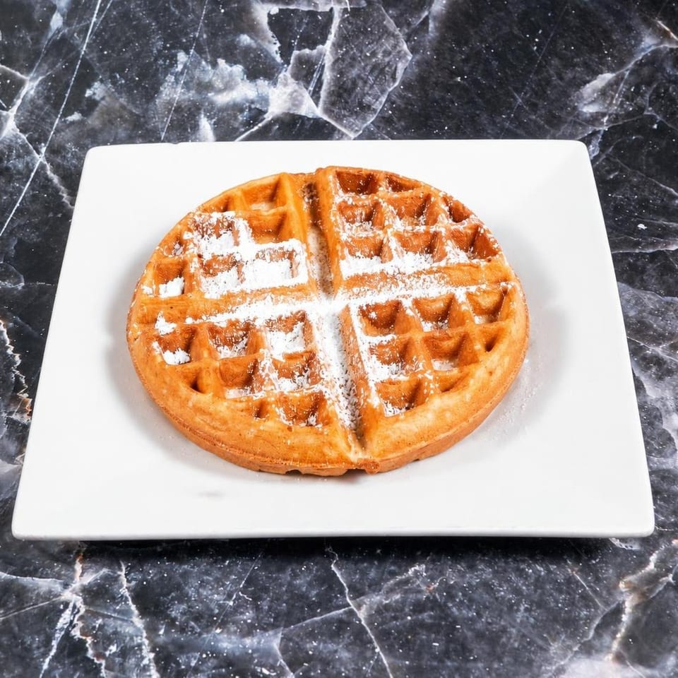 Golden Belgian waffle dusted with powdered sugar on a white plate atop a black marble table.