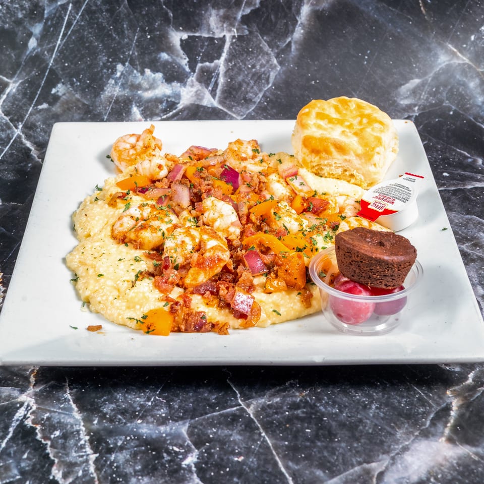 A white plate holds shrimp and grits topped with vegetables and bacon, alongside a biscuit, brownie, fruit, and dipping sauce.