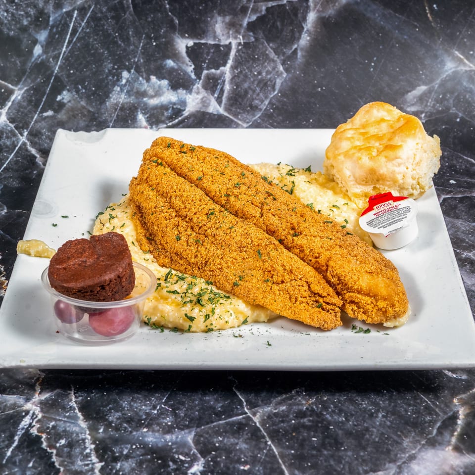 Breaded fried fish fillets served with creamy grits, a biscuit, dipping sauce, and chocolate dessert on a white plate.
